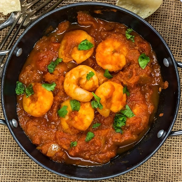 A black pan filled with Indian shrimp curry, garnished with fresh cilantro, on a burlap surface.