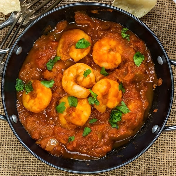 A black pan filled with vibrant red Indian shrimp curry, garnished with fresh green herbs.
